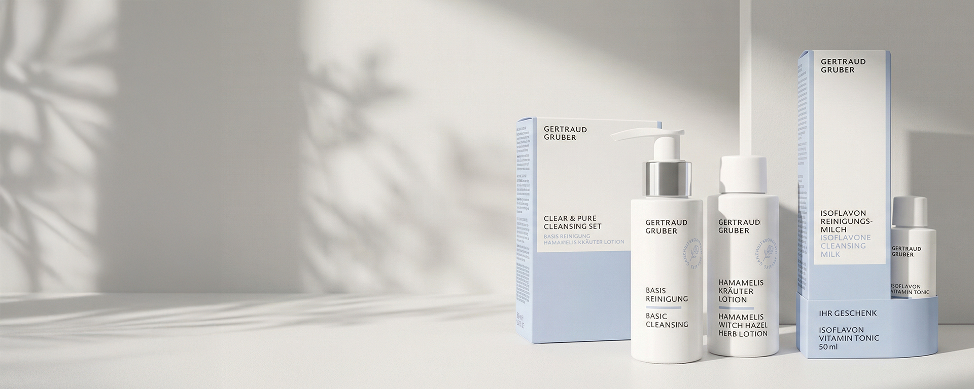 Eine Reihe von weißen und hellblauen Hautpflegeprodukten, einschließlich Flaschen und Schachteln, mit der Aufschrift Gertraud Gruber, ausgestellt auf einer weißen Fläche mit weichen Schatten
